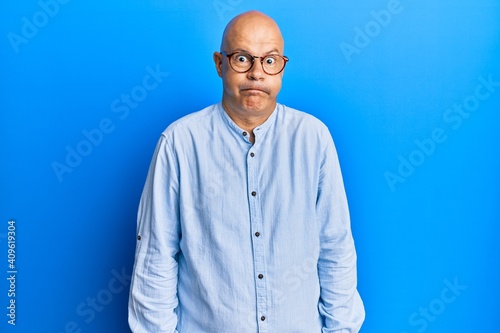Middle age bald man wearing casual clothes and glasses puffing cheeks with funny face. mouth inflated with air, crazy expression.