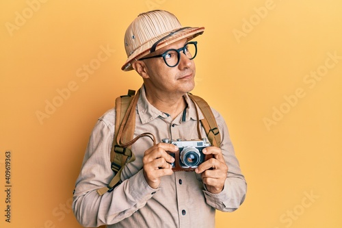 Middle age bald man wearing explorer hat and vintage camera smiling looking to the side and staring away thinking.