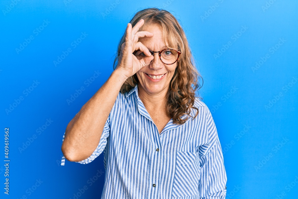 Middle age blonde woman wearing casual clothes and glasses doing ok gesture with hand smiling, eye looking through fingers with happy face. Middle age blonde woman wearing casual clothes and glasses doing ok gesture with hand smiling, eye looking through fingers with happy face.