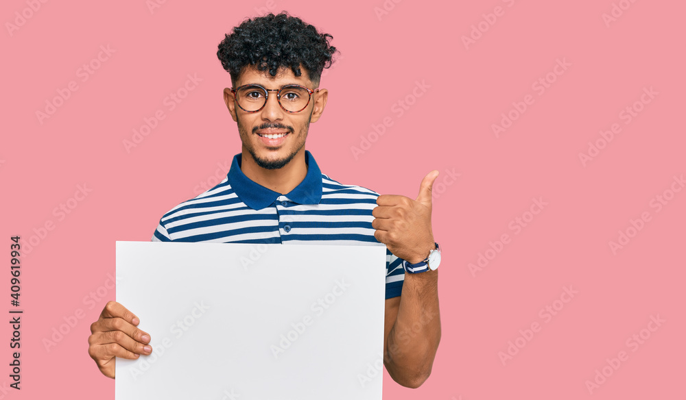Young arab man holding blank empty banner smiling happy and positive, thumb up doing excellent and approval sign Young arab man holding blank empty banner smiling happy and positive, thumb up doing excellent and approval sign