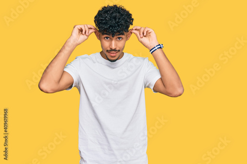 Young arab man wearing casual white t shirt smiling pulling ears with fingers  funny gesture. audition problem