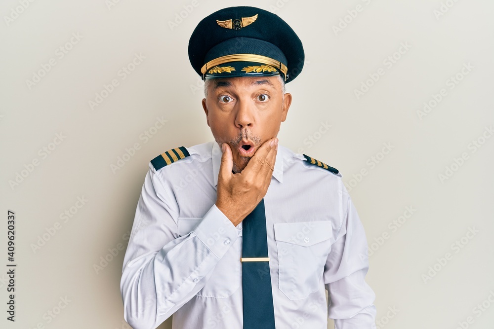 Handsome middle age mature man wearing airplane pilot uniform looking fascinated with disbelief, surprise and amazed expression with hands on chin