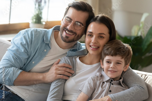 Happy young Caucasian family with little 6s son sit rest on couch in living room hug and cuddle together. Smiling parents relax on sofa at home with small boy child, enjoy lazy leisure weekend.
