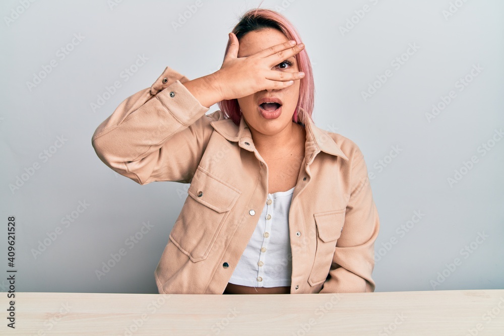 Hispanic woman with pink hair wearing casual clothes sitting on the table peeking in shock covering face and eyes with hand, looking through fingers afraid Hispanic woman with pink hair wearing casual clothes sitting on the table peeking in shock covering face and eyes with hand, looking through fingers afraid