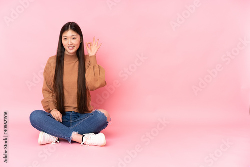 Young asian woman sitting on the floor isolated on pink background showing ok sign with fingers