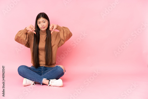Young asian woman sitting on the floor isolated on pink background showing thumb down © luismolinero