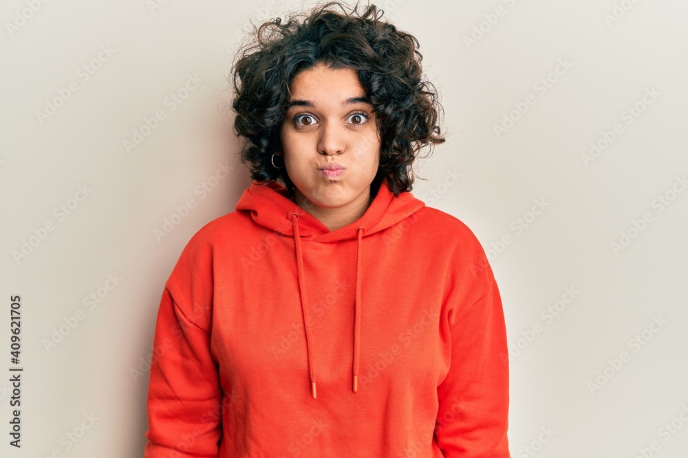 Young hispanic woman with curly hair wearing casual sweatshirt puffing cheeks with funny face. mouth inflated with air, crazy expression.