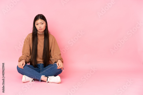 Young asian woman sitting on the floor isolated on pink background with sad and depressed expression