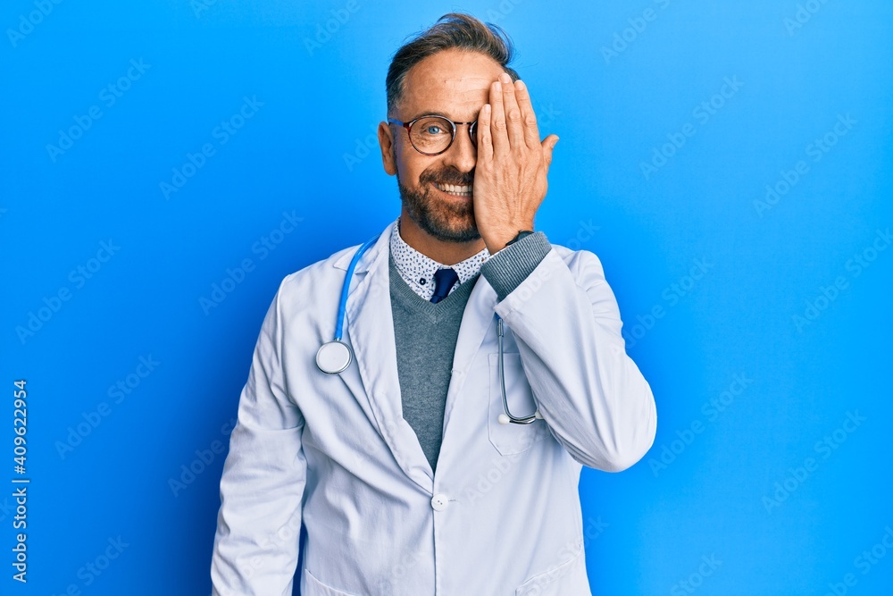 Handsome middle age man wearing doctor uniform and stethoscope covering one eye with hand, confident smile on face and surprise emotion. Handsome middle age man wearing doctor uniform and stethoscope covering one eye with hand, confident smile on face and surprise emotion.