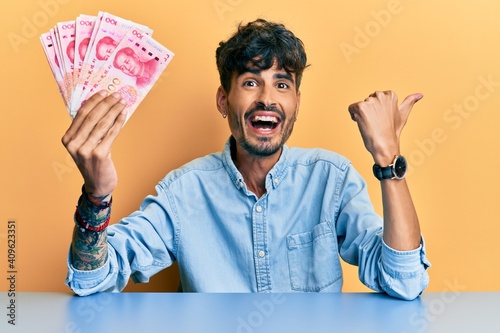 Young hispanic man holding yuan chinese banknotes sitting on the table pointing thumb up to the side smiling happy with open mouth