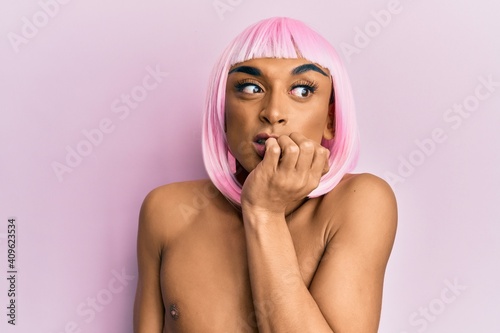 Hispanic transgender man wearing make up and pink wig looking stressed and nervous with hands on mouth biting nails. anxiety problem.