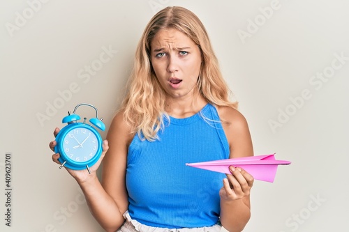 Young blonde girl holding paper plane and alarm clock in shock face  looking skeptical and sarcastic  surprised with open mouth