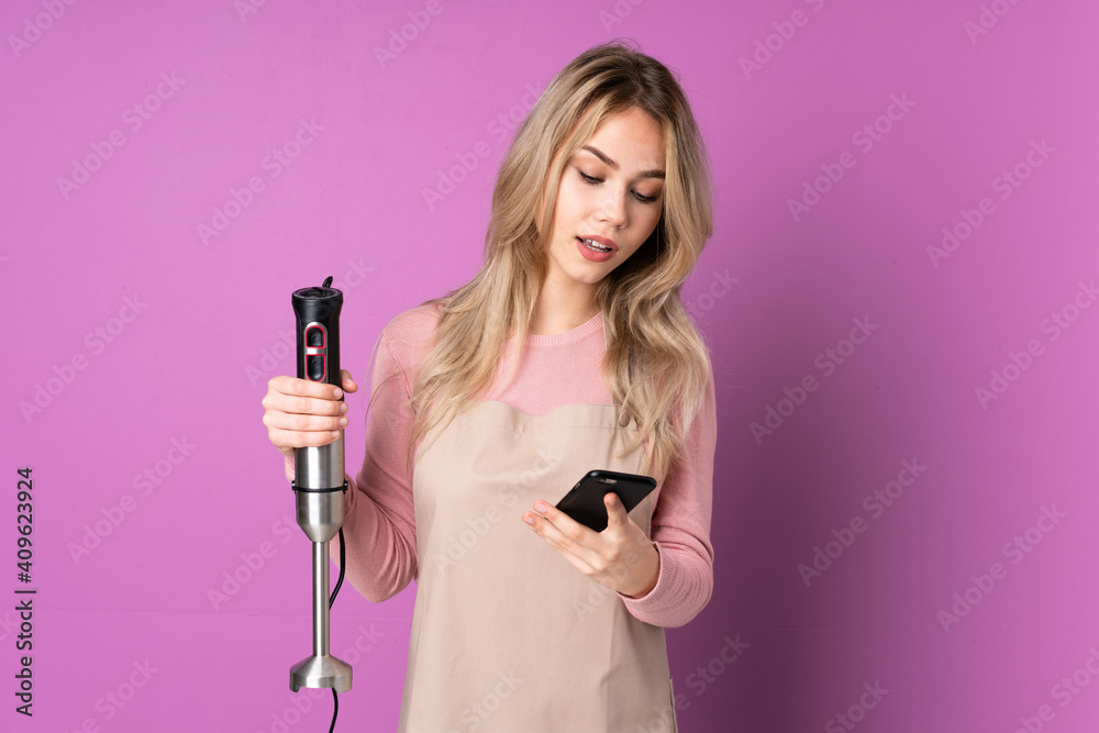 Teenager Russian girl using hand blender isolated on purple background sending a message with the mobile Teenager Russian girl using hand blender isolated on purple background sending a message with the mobile