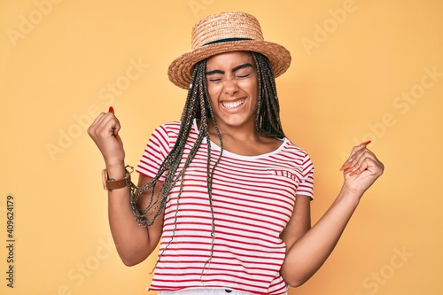 Young african american woman with braids wearing summer hat very happy and excited doing winner gesture with arms raised, smiling and screaming for success. celebration concept.