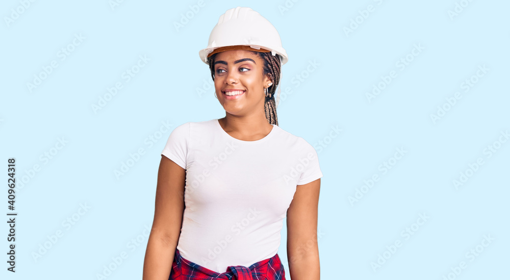 Young african american woman with braids wearing hardhat and builder clothes looking away to side with smile on face, natural expression. laughing confident. Young african american woman with braids wearing hardhat and builder clothes looking away to side with smile on face, natural expression. laughing confident.