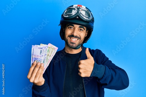 Fototapeta Young hispanic man wearing motorcycle helmet holding indian rupee smiling happy