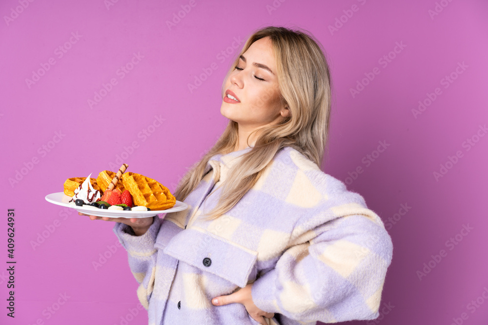 Teenager Russian girl holding waffles isolated on purple background suffering from backache for having made an effort