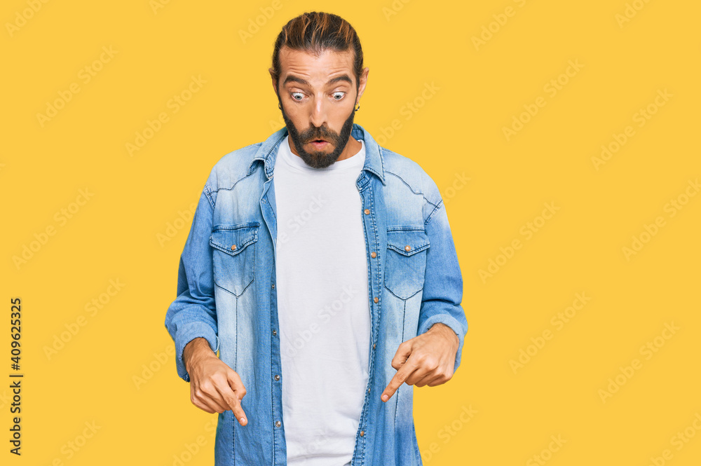 Attractive man with long hair and beard wearing casual denim jacket pointing down with fingers showing advertisement, surprised face and open mouth Attractive man with long hair and beard wearing casual denim jacket pointing down with fingers showing advertisement, surprised face and open mouth