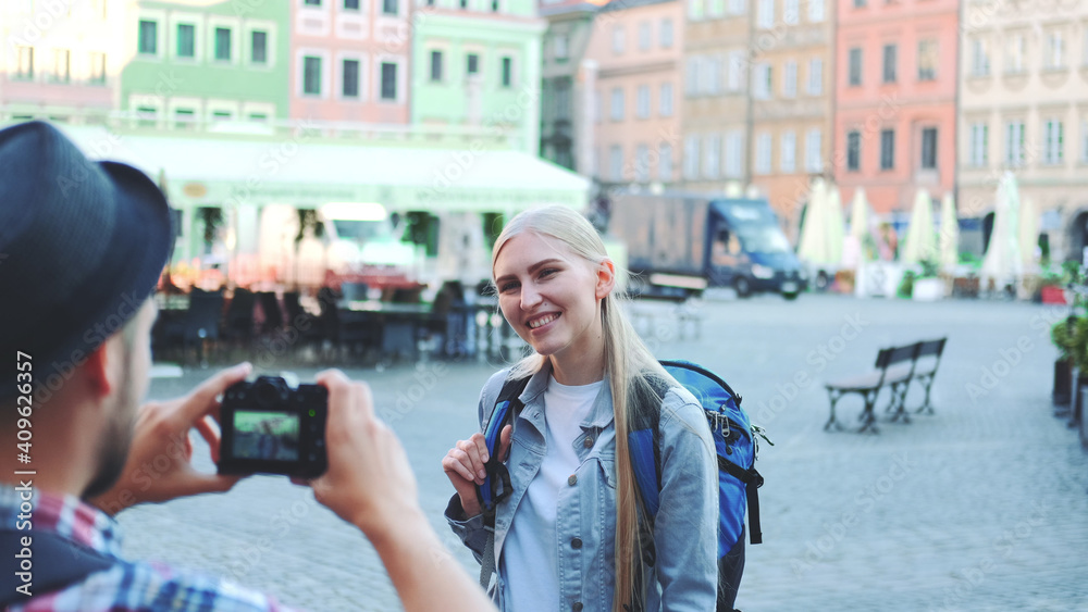 Medium shot of man making photos of female tourist on the view of city market square. Blonde woman making faces and smiling.