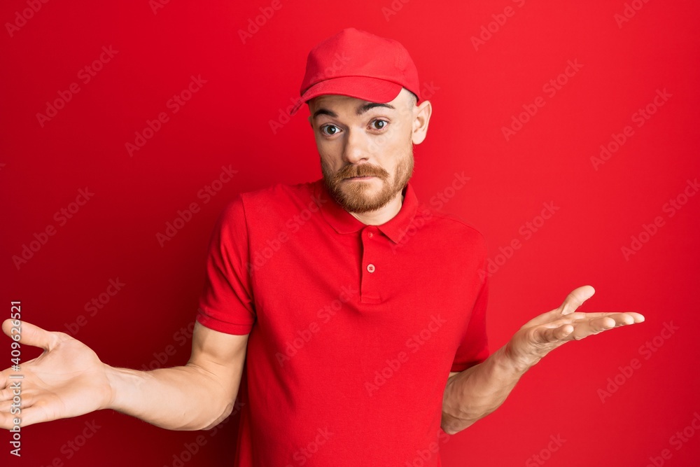 Young redhead man wearing delivery uniform and cap clueless and confused expression with arms and hands raised. doubt concept.