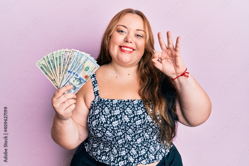 Plus size caucasian young woman holding dollars doing ok sign with fingers, smiling friendly gesturing excellent symbol Plus size caucasian young woman holding dollars doing ok sign with fingers, smiling friendly gesturing excellent symbol