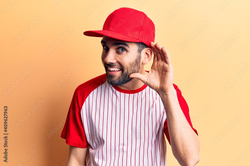 Young handsome man with beard wearing baseball cap and t-shirt smiling with hand over ear listening and hearing to rumor or gossip. deafness concept.