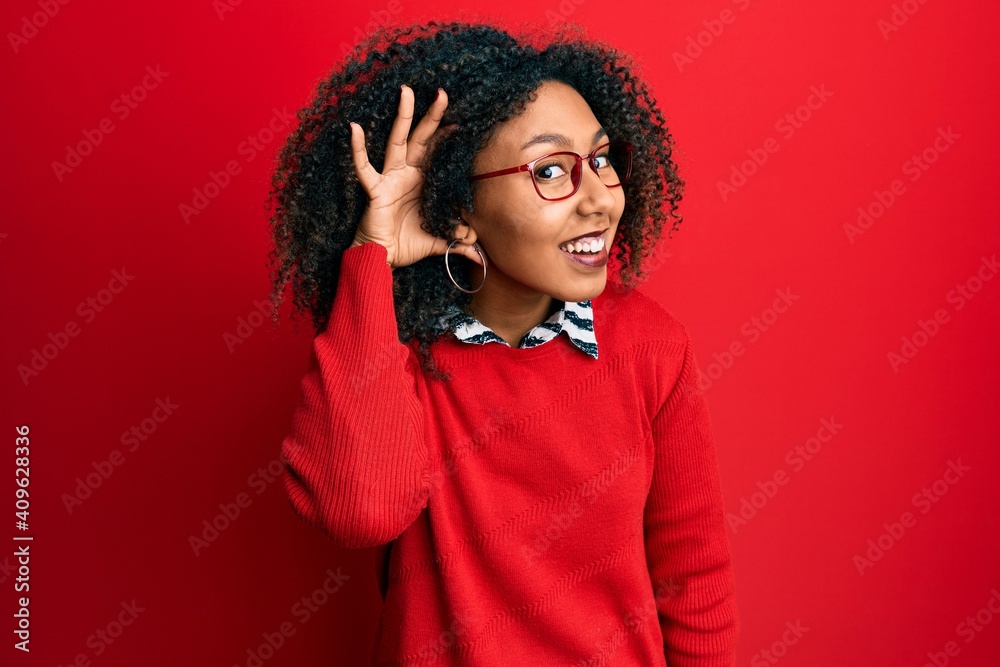 Beautiful african american woman with afro hair wearing sweater and glasses smiling with hand over ear listening an hearing to rumor or gossip. deafness concept. Beautiful african american woman with afro hair wearing sweater and glasses smiling with hand over ear listening an hearing to rumor or gossip. deafness concept.