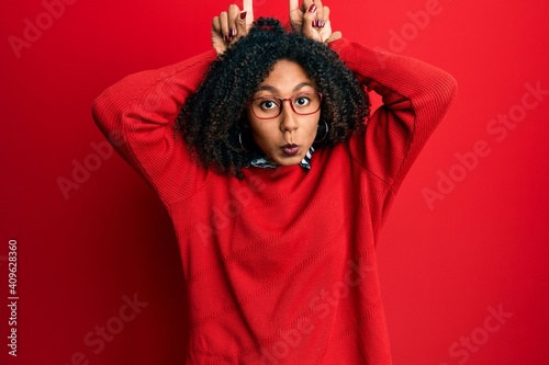 Beautiful african american woman with afro hair wearing sweater and glasses doing funny gesture with finger over head as bull horns © Krakenimages.com