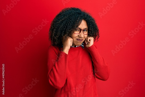 Beautiful african american woman with afro hair wearing sweater and glasses covering ears with fingers with annoyed expression for the noise of loud music. deaf concept. © Krakenimages.com