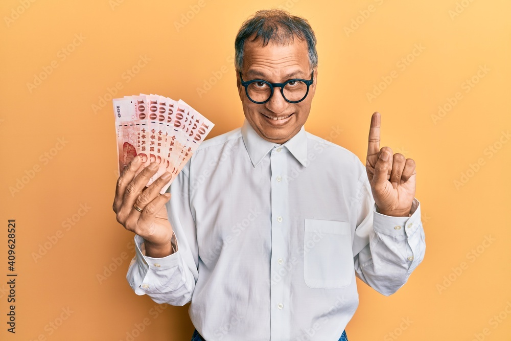 Middle age indian man holding 100 new taiwan dollars banknotes smiling with an idea or question pointing finger with happy face, number one Middle age indian man holding 100 new taiwan dollars banknotes smiling with an idea or question pointing finger with happy face, number one