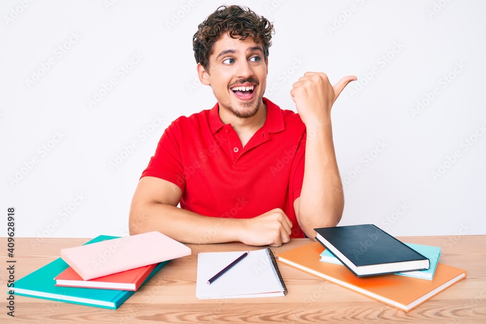 Young caucasian man with curly hair sitting on the table stuying for university pointing thumb up to the side smiling happy with open mouth Young caucasian man with curly hair sitting on the table stuying for university pointing thumb up to the side smiling happy with open mouth