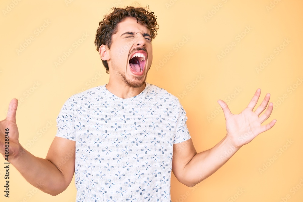 Young caucasian man with curly hair wearing casual clothes crazy and mad shouting and yelling with aggressive expression and arms raised. frustration concept. Young caucasian man with curly hair wearing casual clothes crazy and mad shouting and yelling with aggressive expression and arms raised. frustration concept.