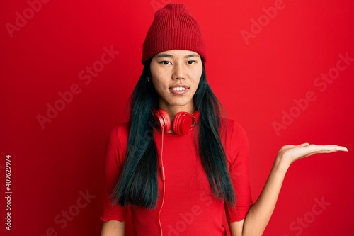 Young chinese woman listening to music using headphones smiling cheerful presenting and pointing with palm of hand looking at the camera.