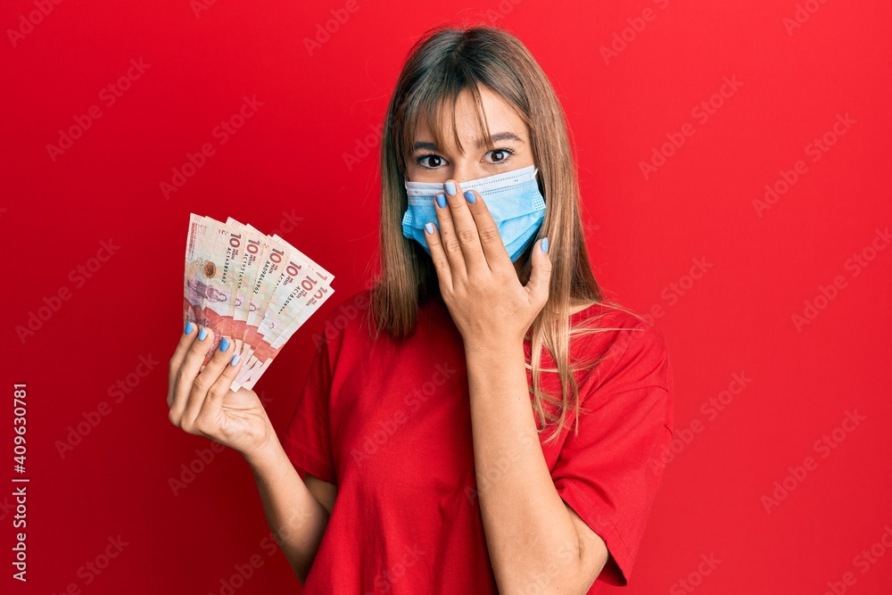 Teenager caucasian girl wearing medical mask holding 10 colombian pesos banknotes covering mouth with hand, shocked and afraid for mistake. surprised expression Teenager caucasian girl wearing medical mask holding 10 colombian pesos banknotes covering mouth with hand, shocked and afraid for mistake. surprised expression