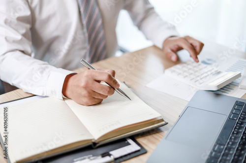 A male employee is taking notes in his notebook, A male employee is using a calculator and taking notes in a notebook.