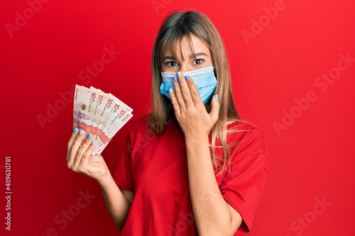 Teenager caucasian girl wearing medical mask holding 10 colombian pesos banknotes covering mouth with hand, shocked and afraid for mistake. surprised expression