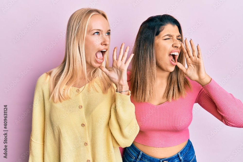 Two friends standing together over pink background shouting and screaming loud to side with hand on mouth. communication concept. Two friends standing together over pink background shouting and screaming loud to side with hand on mouth. communication concept.