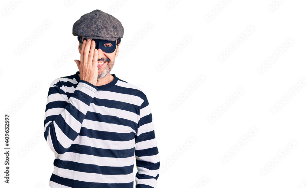 Middle age handsome man wearing burglar mask covering one eye with hand, confident smile on face and surprise emotion. Middle age handsome man wearing burglar mask covering one eye with hand, confident smile on face and surprise emotion.
