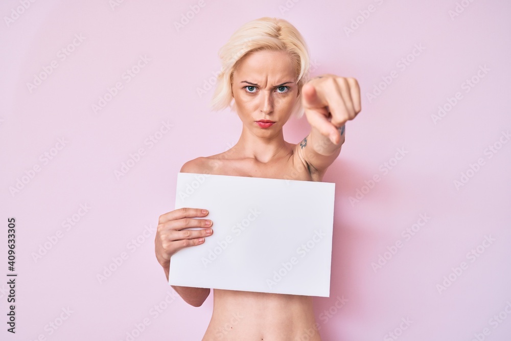 Young blonde woman with tattoo standing shirtless holding empty blank banner pointing with finger to the camera and to you, confident gesture looking serious Young blonde woman with tattoo standing shirtless holding empty blank banner pointing with finger to the camera and to you, confident gesture looking serious