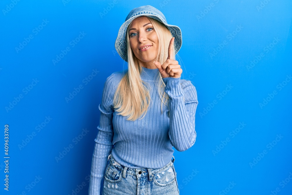 Young blonde woman wearing casual denim hat showing and pointing up with finger number one while smiling confident and happy. Young blonde woman wearing casual denim hat showing and pointing up with finger number one while smiling confident and happy.