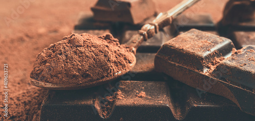 Brown cocoa powder in the spoon and chopped chocolate cubes, on dark background