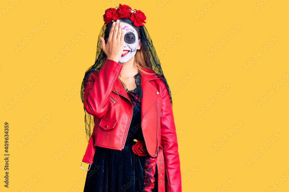 Woman wearing day of the dead costume over background covering one eye with hand, confident smile on face and surprise emotion. Woman wearing day of the dead costume over background covering one eye with hand, confident smile on face and surprise emotion.