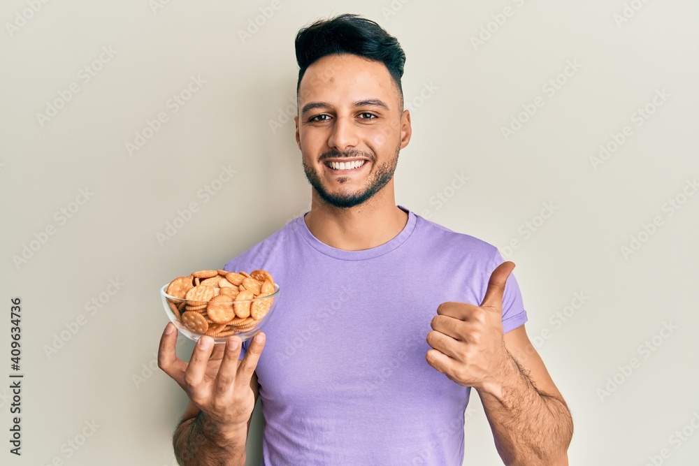 Young arab man holding bowl of salty crackers biscuits smiling happy and positive, thumb up doing excellent and approval sign Young arab man holding bowl of salty crackers biscuits smiling happy and positive, thumb up doing excellent and approval sign