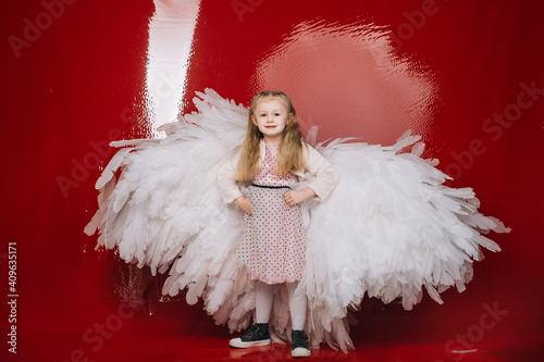 little girl 4 years old in white angel wings on a red latex background in the studio for valentine's day