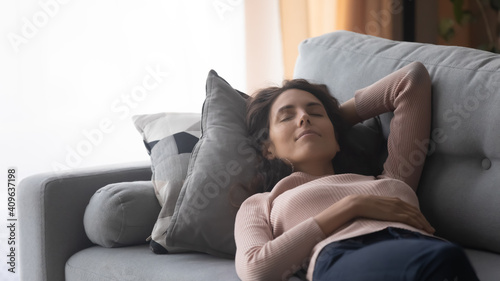 Relaxed millennial woman sleeping on comfortable sofa, daydreaming alone at home, enjoying sweet dreams on lazy weekend. Happy young lady lying on couch with close eyes, meditating or napping.
