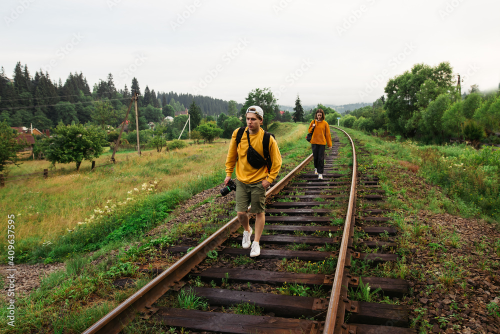 Couple of tourists with backpacks walking on the railway track in the mountains. Woman man hipsters walking in the mountains on rails.