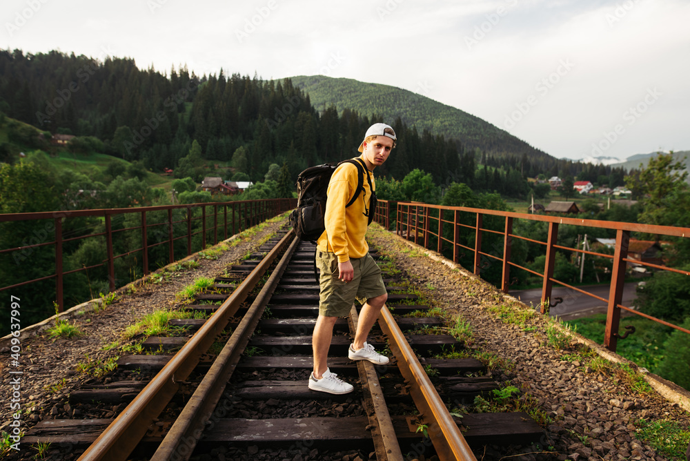 A guy in an orange sweater stands on a track on a railway bridge in the mountains and looks at the camera. young male tourist walks on a viaduct in the mountains