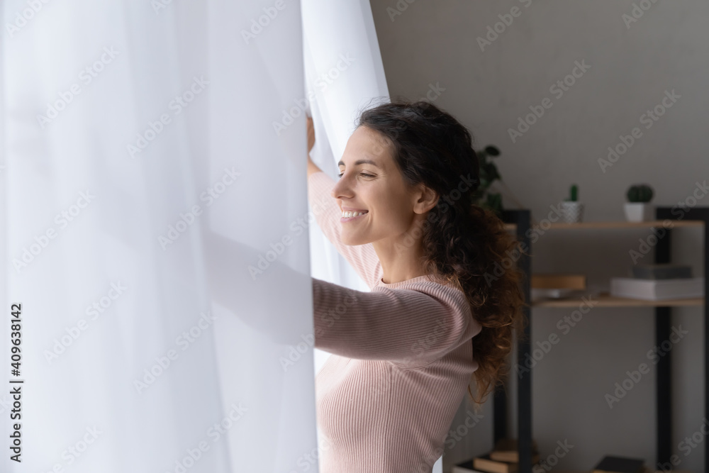 Side view happy young beautiful caucasian woman opening curtains, enjoying morning cityscape view, welcoming new sunny day, feeling motivated inspired alone at home or hotel, dreaming indoors. Side view happy young beautiful caucasian woman opening curtains, enjoying morning cityscape view, welcoming new sunny day, feeling motivated inspired alone at home or hotel, dreaming indoors.