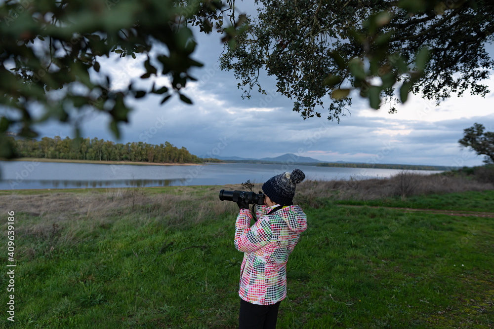Photo of a young and attractive female wearing winter clothes and taking pictures of wildlife close to a lake during the winter season.