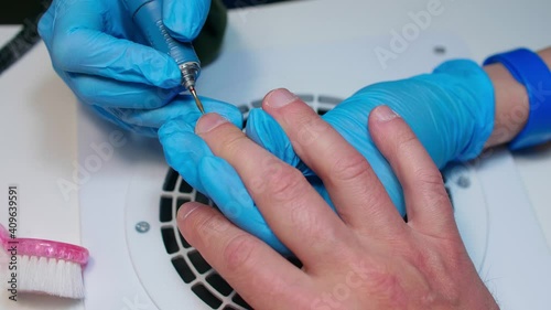 Manicurist in blue gloves removes dry skin around nail with machine nailfile on man's hand. Close up view. photo